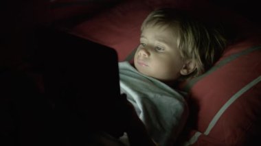 Sarı saçlı genç bir çocuk, tablet ekranına odaklanmış, gece karanlığında yüzünü aydınlatan cihazlar..