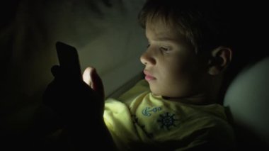 Kahverengi saçlı bir çocuk, tablet bir ekrana odaklanmış, gece karanlık bir odada yüzünü aydınlatan cihazlar..