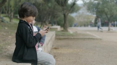Akıllı telefonlu genç bir çocuk, parkta bir bankta öylesine oturuyor ve yanında sakin bir açık hava ortamında sohbet ediyor gibi görünüyor..