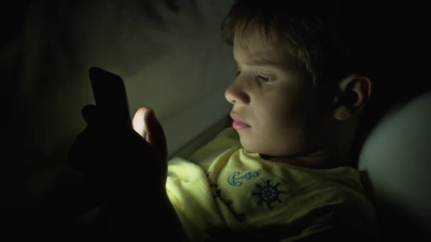 茶色の髪の少年が下に横たわり タブレットの画面に焦点を当て デバイスは夜の暗い部屋で顔を明るく照らしています — ストック動画