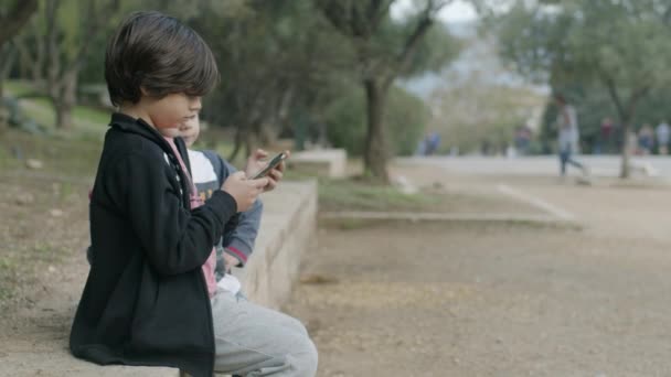 スマートフォンを持った少年が公園のベンチに気軽に座り 静かな屋外環境の中で隣の誰かと会話しているようだ — ストック動画