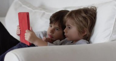 Çocuklar oyuncakları keşfediyor ve öğreniyor, birlikte eğlenceli bir aktiviteye giriyorlar, bu sırada parlak, modern bir oturma odasında dijital tablet kullanıyorlar..