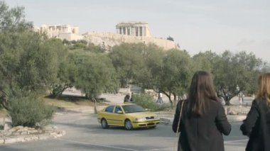Huzurlu bir manzara, zeytin ağaçlarının yanında gezinen ziyaretçileri ve arka planda açık bir gökyüzünün altında yükselen antik Akropolis 'i yakalıyor..