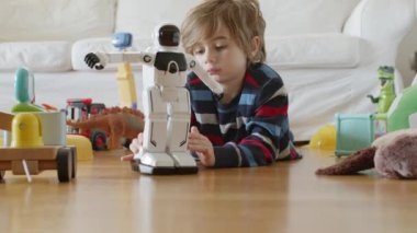 Sarı saçlı genç bir çocuk, dağınık oyuncakların arasında beyaz bir robot oyuncakla oynamaya odaklanmış..