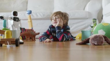 Sarı saçlı genç bir çocuk, dağınık oyuncakların arasında beyaz bir robot oyuncakla oynamaya odaklanmış..