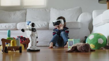 Bir çocuk, oturma odasında VR gözlükleriyle yerde oturuyor, çeşitli oyuncaklarla dolu bir robot oyuncakla etkileşime geçiyor ve yanında pelüş bir oyuncak var..