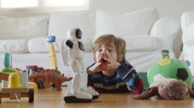 Sarı saçlı genç bir çocuk, dağınık oyuncakların arasında beyaz bir robot oyuncakla oynamaya odaklanmış..