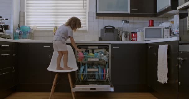 一个男孩站在凳子上走到洗碗机前 小心翼翼地把碗碟放在里面 在厨房里表现出责任感 — 图库视频影像
