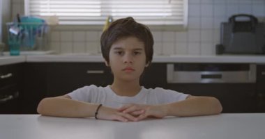 Kahverengi saçlı bir çocuk mutfak masasında oturuyor ve kolları önünde çapraz duruyor. Tarafsız bir ifadeyle dik dik bakıyor..