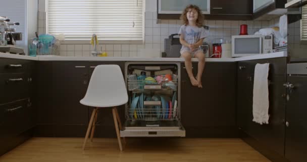 一个留着卷发的男孩坐在厨房的柜台上 他的腿悬在一个盛满碗碟的开放式洗碗机上 准备洗碗 — 图库视频影像