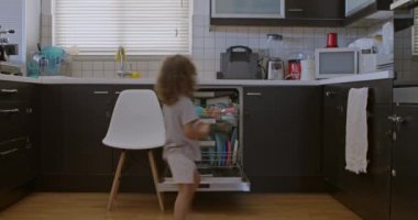 Bir çocuk açık bulaşık makinesine ulaşmak için tabureye çıkıyor. Bulaşıkları dikkatlice içine yerleştiriyor. Mutfak ortamında sorumluluk gösteriyor..