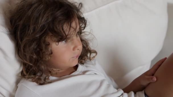 一个卷曲长发的小男孩舒服地躺在沙发上 沉思地凝视着远方 — 图库视频影像