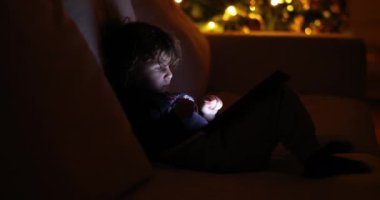 Küçük bir çocuk bilgisayar ekranının önünde oturuyor, dijital tabletle etkileşime girerken odaklanmış ve meşgul..