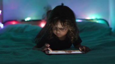 Loş bir odada karnının üstüne uzanmış, arka planda renkli ortam ışıkları olan dijital bir tablete odaklanmış bir çocuk..