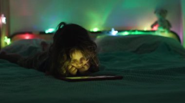 Bir çocuk göbeğinde yatar, dijital tablete odaklanır, gecenin karanlığında çok renkli ışıkların yumuşak parıltısıyla yıkanır..