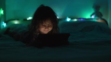 Loş bir odada karnının üstüne uzanmış, arka planda renkli ortam ışıkları olan dijital bir tablete odaklanmış bir çocuk..