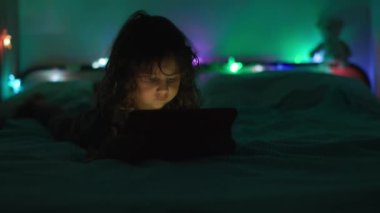 Loş bir odada karnının üstüne uzanmış, arka planda renkli ortam ışıkları olan dijital bir tablete odaklanmış bir çocuk..