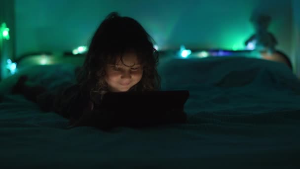 一个小男孩躺在光线昏暗的房间里 专心致志地盯着背景上有五彩缤纷的环境灯的数码平板电脑 — 图库视频影像
