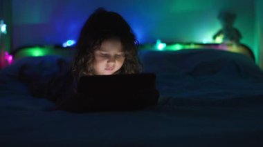 Loş bir odada karnının üstüne uzanmış, arka planda renkli ortam ışıkları olan dijital bir tablete odaklanmış bir çocuk..