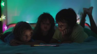 Dijital ekranın parıltısı altında, üç çocuk bir yatağa uzanır, gece boyunca tablette gösterilen içeriğe gömülürler..