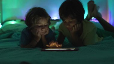 Dijital ekranın parıltısı altında, iki çocuk bir yatağa uzanmış, gece boyunca tablette gösterilen içeriğe gömülmüş..