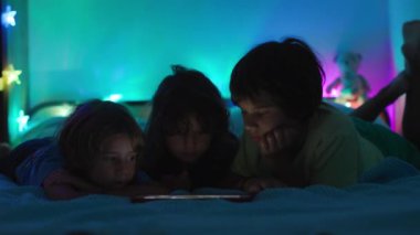 Dijital ekranın parıltısı altında, üç çocuk bir yatağa uzanır, gece boyunca tablette gösterilen içeriğe gömülürler..