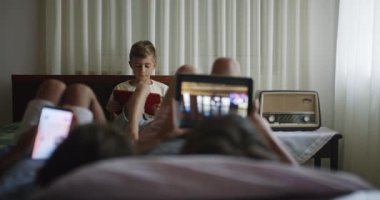 İki erkek çocuk bir yatakta yatarken dijital tablet kullanırken, diğer erkek çocuk arka planda dijital tablet kullanarak rahat ve rahat bir atmosfer yakalıyor..