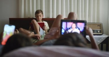 İki erkek çocuk bir yatakta yatarken dijital tablet kullanırken, diğer erkek çocuk arka planda dijital tablet kullanarak rahat ve rahat bir atmosfer yakalıyor..