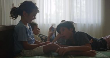 İki çocuk, aralarında dijital tablet bulunan bir yatakta uzanıyorlar. Ev ortamındaki ekran içeriğine derinden odaklanmış durumdalar..