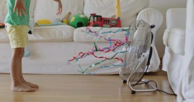 Uzun saçlı neşeli bir çocuk ahşap zeminde oturuyor, elektrikli yelpazeye bağlı renkli kurdelelerle oynuyor, rüzgarı içeride hissediyor..