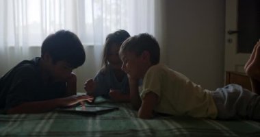 Üç çocuk, aralarında dijital tabletle bir yatakta uzanıyorlar, akşam yaklaşırken loş bir odada birbirleriyle meşgul oluyorlar..