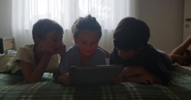 Üç çocuk, aralarında dijital tabletle bir yatakta uzanıyorlar, akşam yaklaşırken loş bir odada birbirleriyle meşgul oluyorlar..