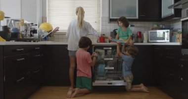 Bir anne, bulaşıkları bulaşık makinesine koymaya yardım ederken çocuklarına göz kulak olur. İşbirliği duygusunu ve aile sorumluluğunu teşvik eder..