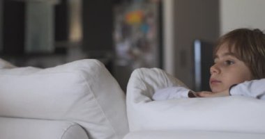 Düşünceli genç bir çocuk, beyaz bir kanepede uzanmış, rüya gibi bakışlarıyla başka yere bakarken, çenesini ellerinin üzerinde dinlendirir..