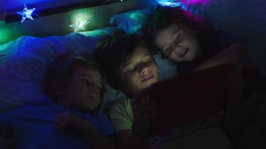 Bir grup çocuk yan yana yatıp dijital bir tablo kullanıyorlar. Ekran, önlerindeki dijital tablete odaklandıklarında yüzlerini aydınlatıyor..