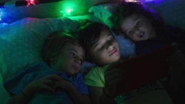 Bir grup çocuk yan yana yatıp dijital bir tablo kullanıyorlar. Ekran, önlerindeki dijital tablete odaklandıklarında yüzlerini aydınlatıyor..