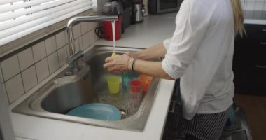 Bir kadın mutfak lavabosunda duruyor, mavi tabakları musluktan suyla duruluyor..