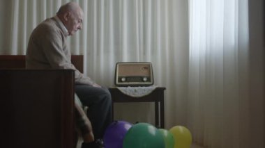Yaşlı bir beyefendi, canlı balonlarla çevrili, bir radyonun yanındaki sandalyeye oturmuş, kutlama ve yalnızlığın yan yana geldiğini öne sürüyor..