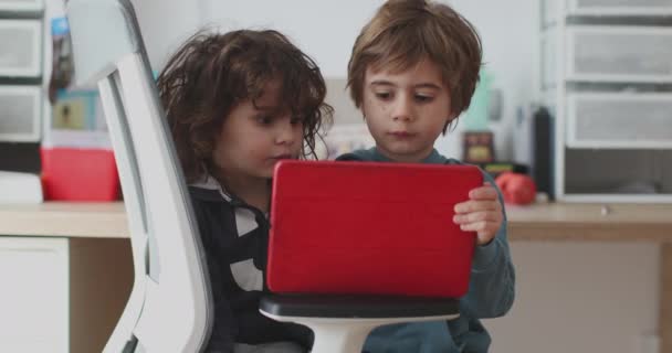 2人の小さな少年がデジタルタブレットに焦点を当てながら密接に座り 子供の寝室を示す背景にはおもちゃと本棚がありました — ストック動画