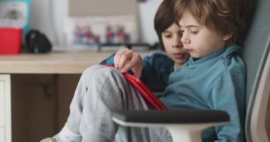 İki küçük çocuk, dijital tablete dikkatle odaklanırken, arka planda bir kitaplık ve gün boyunca bir çocuğun yatak odasını gösteren oyuncaklarla birlikte oturuyorlar..
