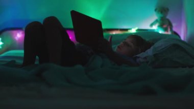 Genç bir çocuk loş bir odada uzanıyor, arka planda renkli ortam ışıkları olan dijital bir tablete odaklanıyor..