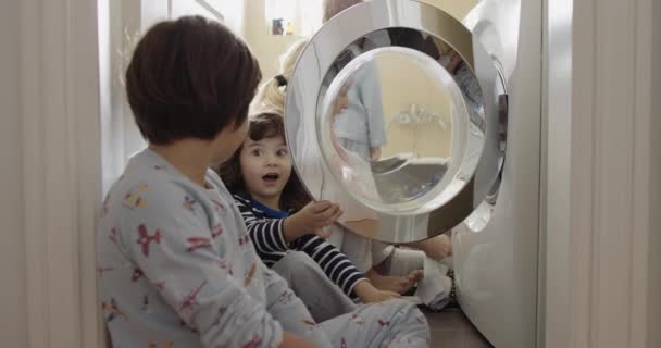 一位母亲照看着她穿着睡衣的小孩 他们看着衣服在洗衣机里旋转 把家务活粘在一起 — 图库视频影像