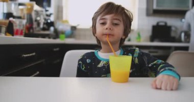 Genç bir çocuk pipetle portakal suyunu yudumluyor, uzay temalı bir gömlek giyiyor, mutfak masasında oturuyor ve gün ışığı içeri giriyor..