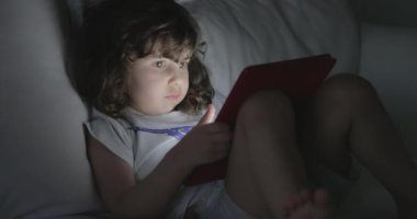 Genç bir çocuk beyaz bir kanepede rahatça oturur. Dikkatini, sakin alacakaranlık saatlerinde elinde tuttuğu kırmızı tablet çeker..