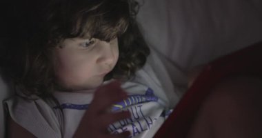 Genç bir çocuk beyaz bir kanepede rahatça oturur. Dikkatini, sakin alacakaranlık saatlerinde elinde tuttuğu kırmızı tablet çeker..