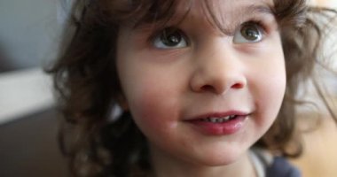 Kıvırcık saçlı ve mavi gömlekli genç bir çocuk mutfakta dikilip, tarafsız bir ifadeyle izleyiciye dikkatlice bakıyor..