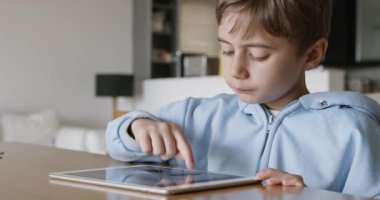 Sarı saçlı genç bir çocuk, gün boyunca, iyi aydınlatılmış, ferah bir oturma odasında, ahşap bir masada otururken dijital tablet kullanmaya odaklanır..