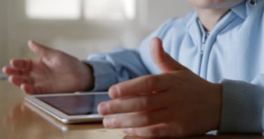 Sarı saçlı genç bir çocuk, gün boyunca, iyi aydınlatılmış, ferah bir oturma odasında, ahşap bir masada otururken dijital tablet kullanmaya odaklanır..