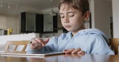 Sarı saçlı genç bir çocuk, gün boyunca, iyi aydınlatılmış, ferah bir oturma odasında, ahşap bir masada otururken dijital tablet kullanmaya odaklanır..