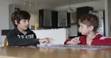 Genç oğlanlardan oluşan odaklanmış bir ikili, içeride ahşap bir masa üzerinde canlı bir yapboz parçalarını bir araya getirerek problem çözme becerilerini sergiliyor..